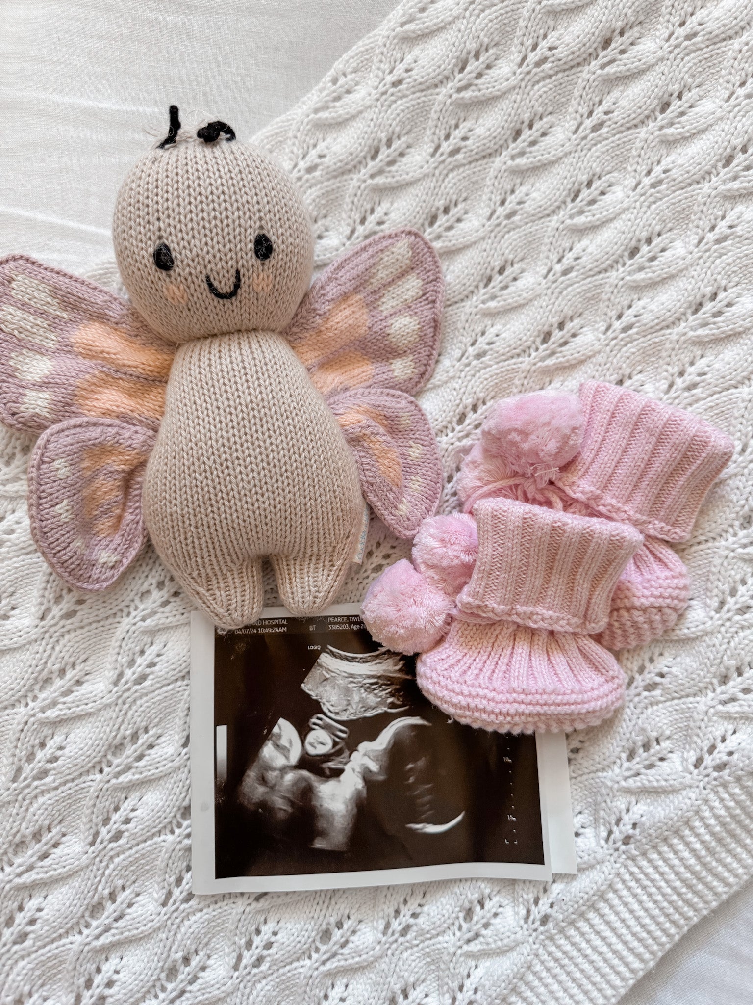 Little B's Knitted Pom Pom Booties in pink, a knitted butterfly toy, and a sonogram on a white blanket