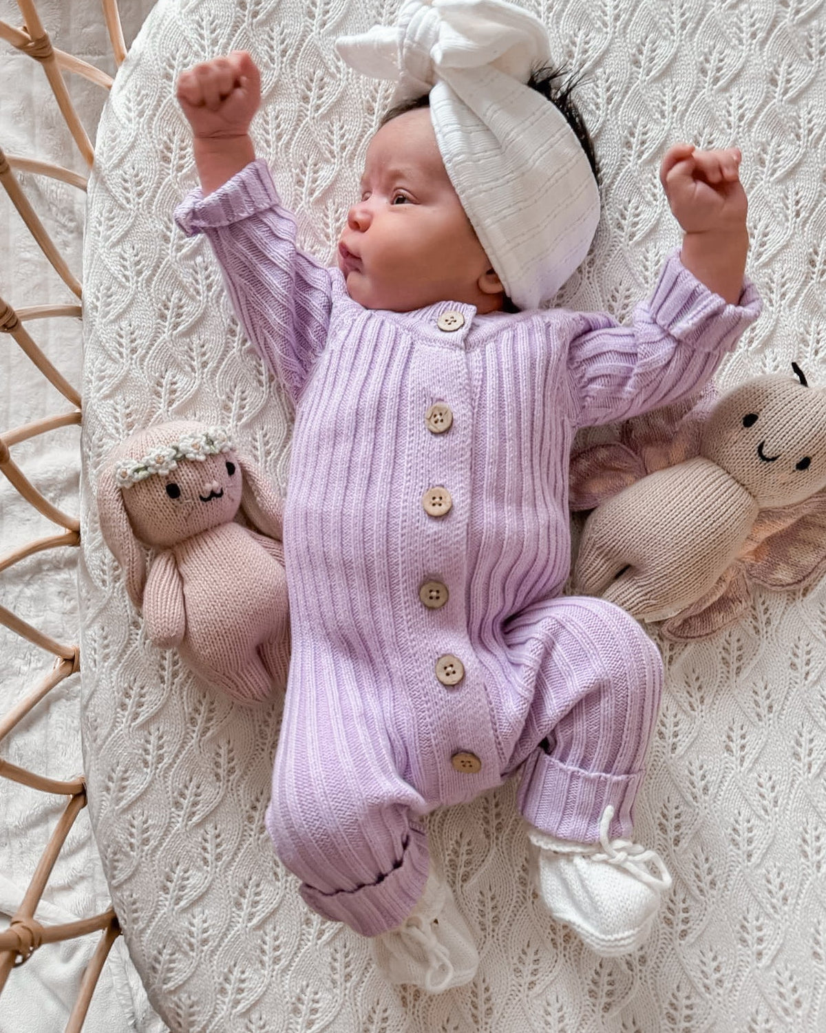 A baby lying on a textured cream knit blanket, wearing a light purple ribbed long-sleeve footless romper with wooden buttons, a white bow headband, and white booties. Two knitted stuffed sheep are on either side of the baby.