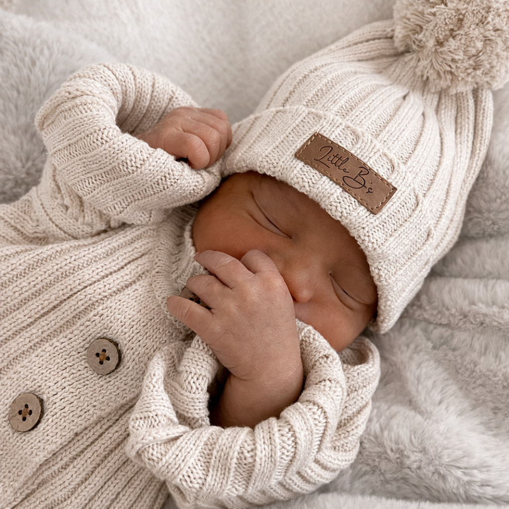 Newborn baby sleeping in a cream knitted Little B's outfit with a pom-pom hat