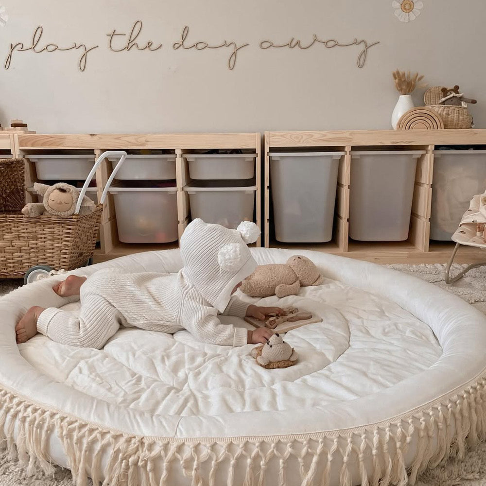 Baby in white knitted outfit with pom-pom hat playing on a round white play mat with fringe